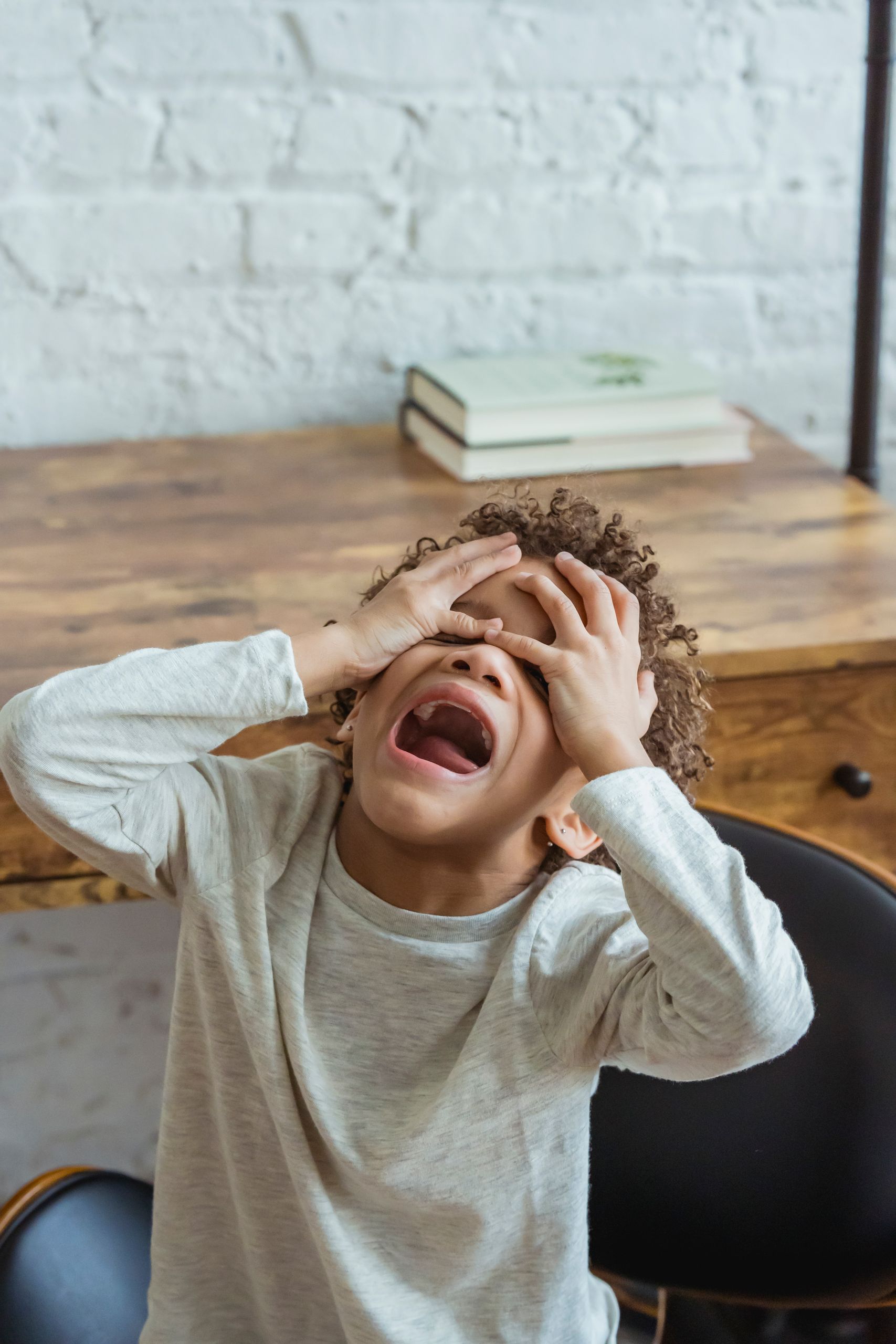 Unhappy African American boy with curly hair in casual wear covering face with hands while shouting in room at home